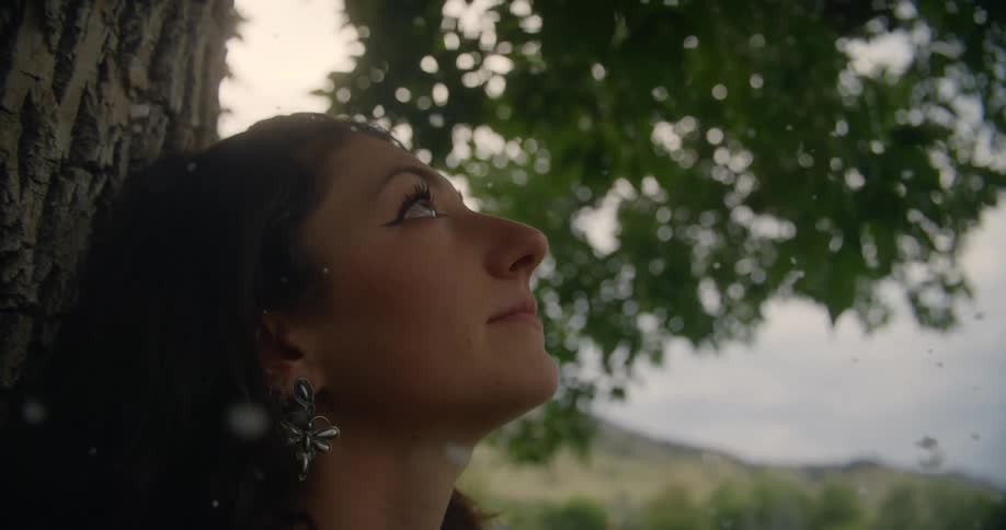 A woman with long dark hair and a flowershaped earring leans against a tree trunk surrounded by white motes with green leaves and a distant landscape in the background