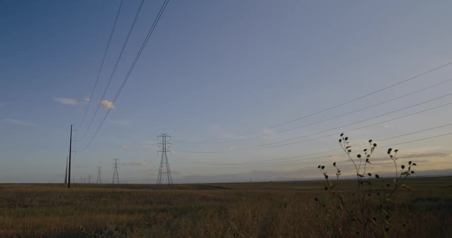 A vast dry field stretches under a blue sky dotted with fluffy white clouds bisected by tall electrical pylons and their connecting wires