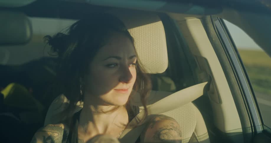 A tattooed woman with hair flowing in the wind drives a car through a rural landscape on a sunny day