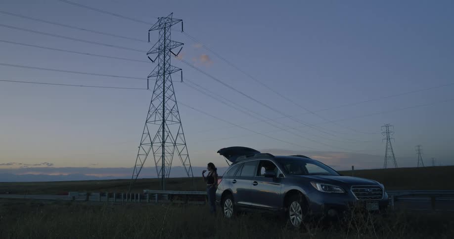 A woman stands next to a parked suv with the trunk open brushing her teeth under a large power transmission tower in a rural area during dusk