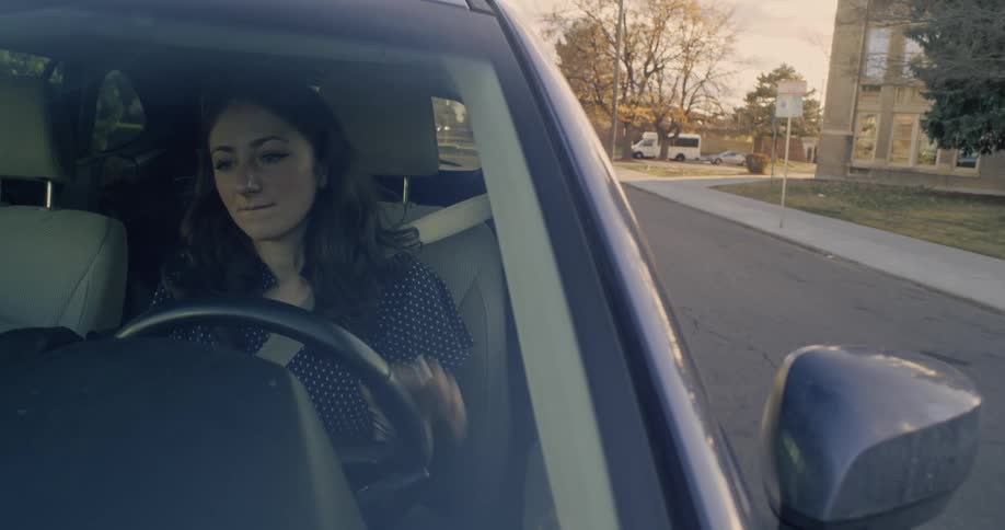 A woman drives a car backing out of the driveway and then onto city streets in the evening