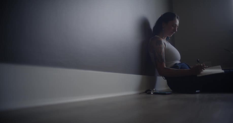 A woman with a tattooed arm sits on the floor in a dimly lit room writing in a notebook