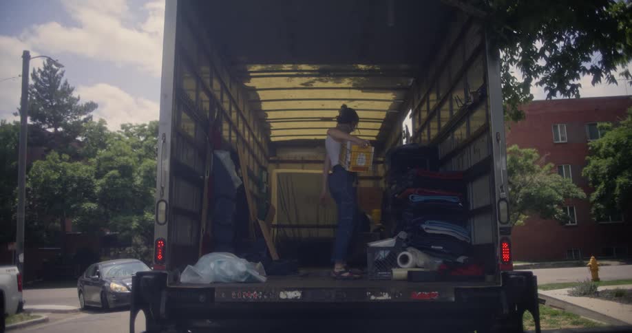 A person is standing inside the back of a moving truck organizing items and holding a basket with various packing materials and blankets visible around