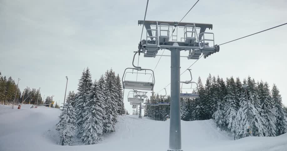 A chairlift ascends a snowy slope carrying empty chairs towards a line of evergreen trees bordering the edge of a mountain under a cloudy sky
