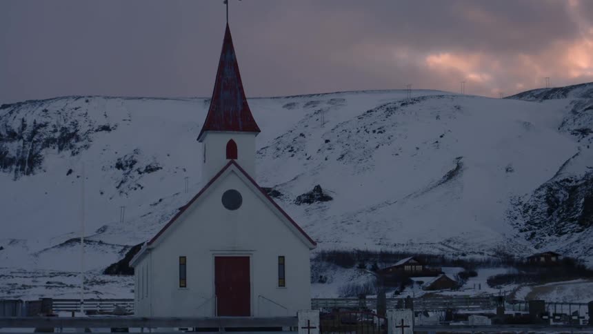 A small white church with a red roof and steeple stands in a snowy mountainous landscape under a cloudy sky