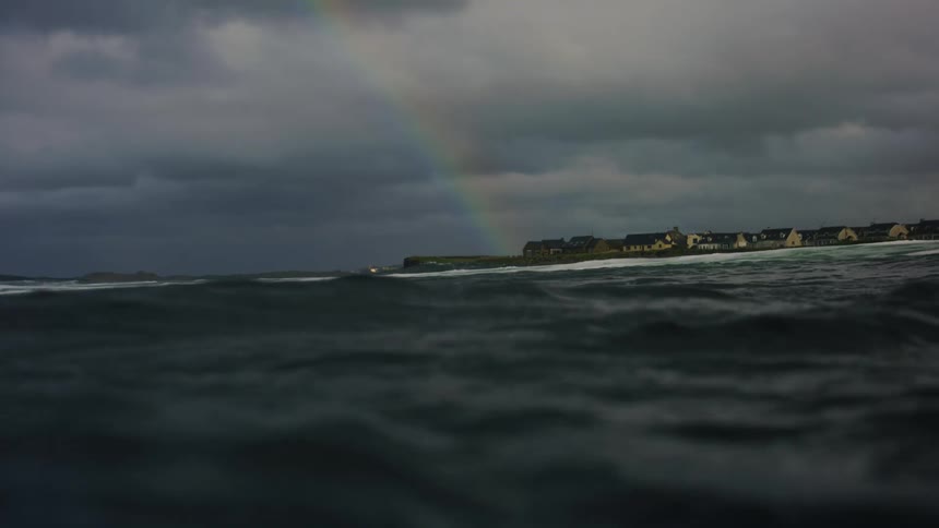 A sea with dark water and some waves coming from the left and a colorful rainbow in a gray sky