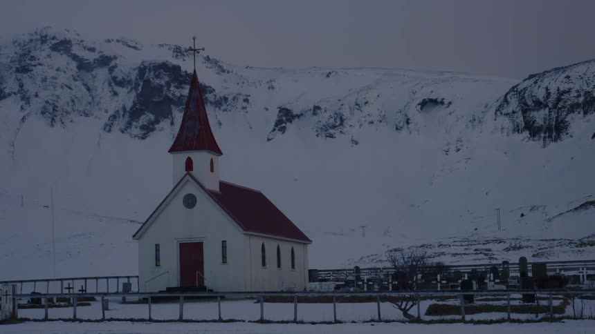 A small white church with a red roof and steeple stands in a snowy mountainous landscape under a cloudy sky