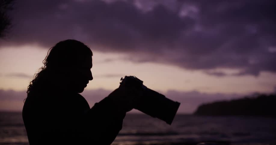 Silhouetted photographer on the shore takes a picture with his camera after dusk