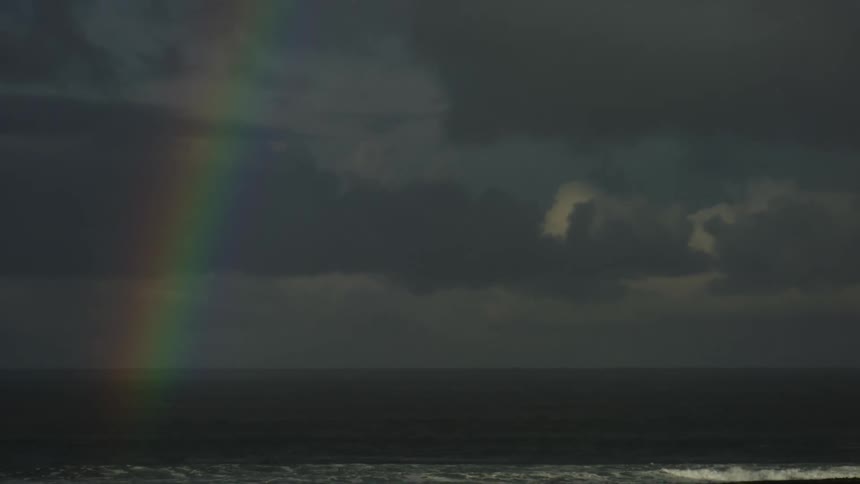 A vibrant rainbow stretches across a dark cloudy sky above the calm ocean creating a serene contrast as sunlight breaks through the stormy clouds