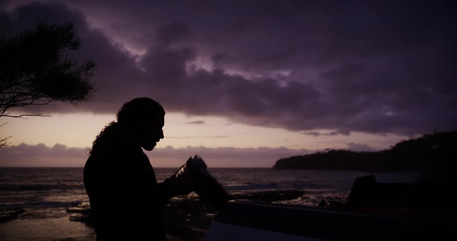 A silhouette of a person with curly hair takes a photograph with a large camera lens at sunset by the ocean with dramatic clouds and a distant shoreline in the background