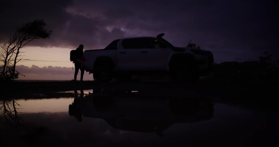 Silhouetted against a dusky sky a person stands next to a parked pickup truck near a reflective body of water