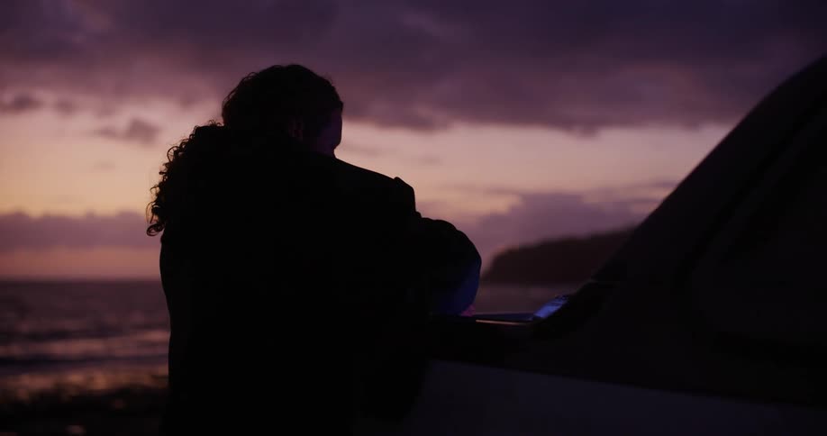 A silhouette of a person picking up a piece of clothing from behind a car on a dark morning