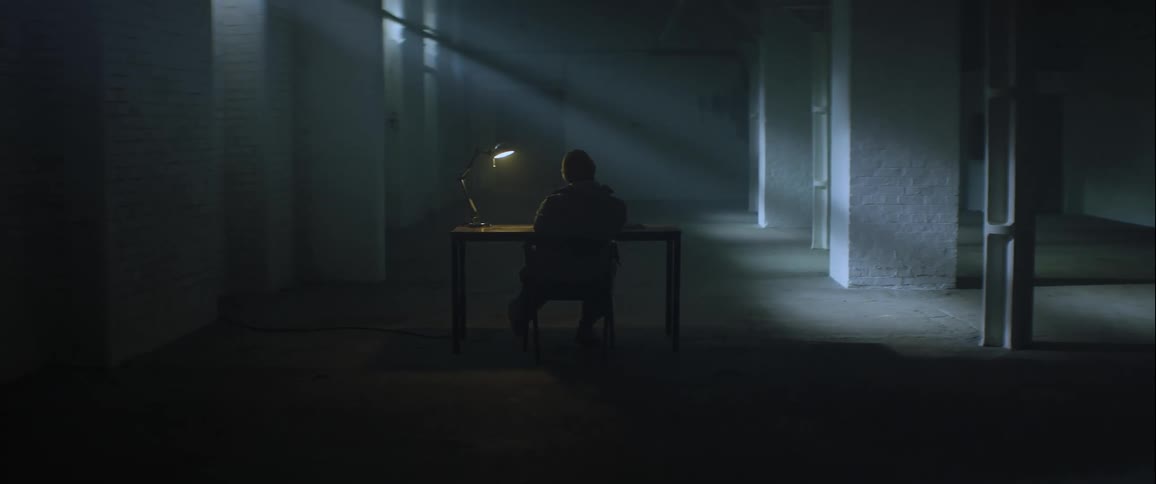 A lone figure sits at a desk illuminated by a lamp in a dimly lit cavernous room with rows of pillars