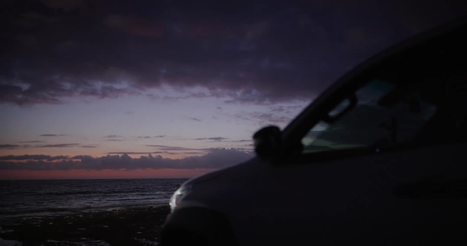 A white suv is parked along a dark beach at dusk facing the ocean under a dramatic cloudy sky with a faint glow on the horizon