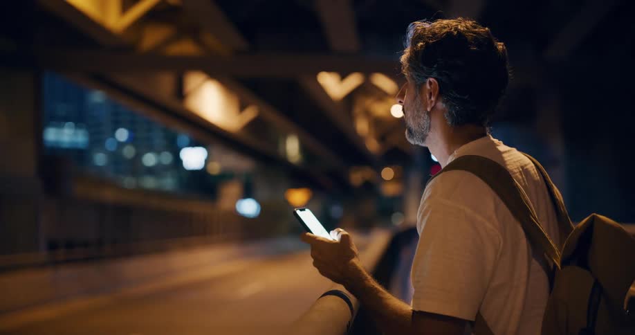 A man stands on an urban bridge at night focused on his iphone with soft city lights and blurred buildings in the background