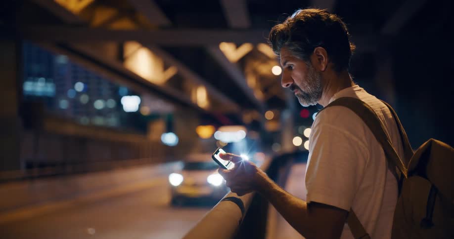 Man standing on a pedestrian path near the highway watches traffic while using his smartphone