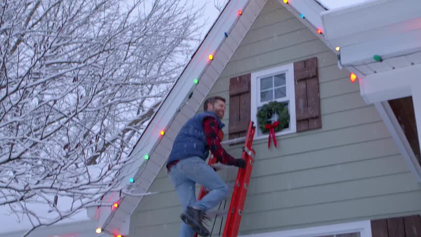 Man uses a ladder to decorate his house with christmas lights and christmas wreaths during a snowfall