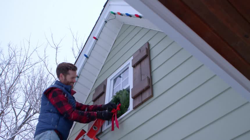 Man placing a ladder outside his house and climbing up it to adjust christmas decorations