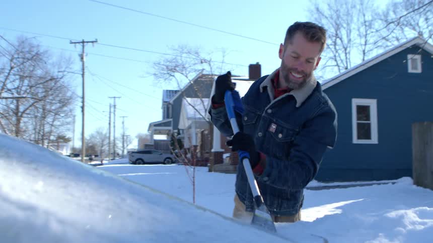 A man scrapes ice from the windshield of a car parked in a neighborhood on a sunny winter morning