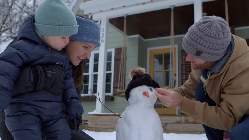 Dad adding eyes to snowman as mom and baby watch