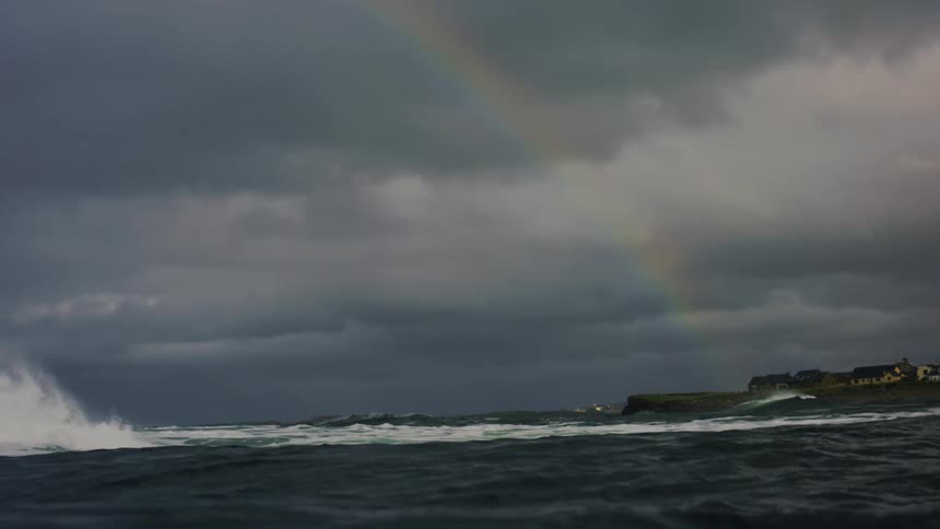 Rainbow arcs across the dark clouds near a coastal town while large waves flow across the sea