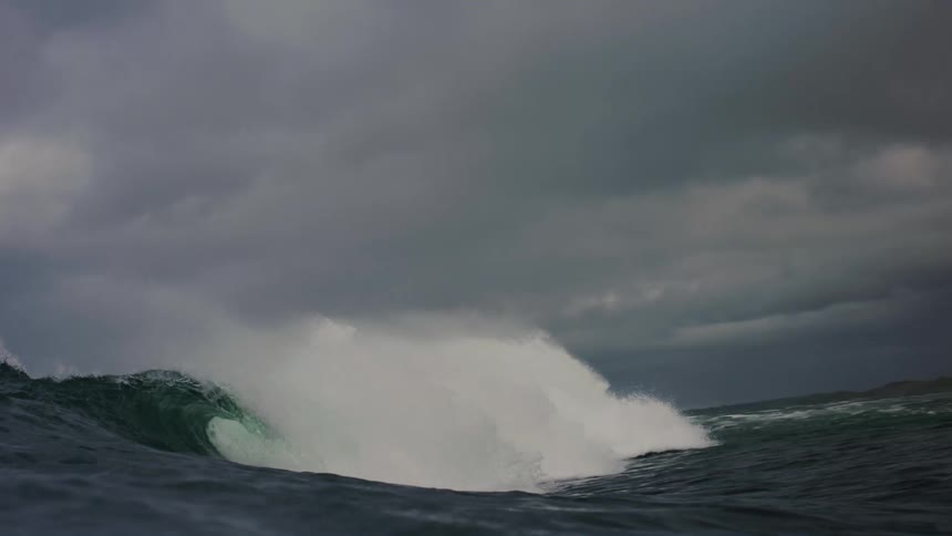 A vibrant rainbow arches across a stormy sky above a churning ocean where a wave crashes against a rocky breakwater sending up a spray of white foam