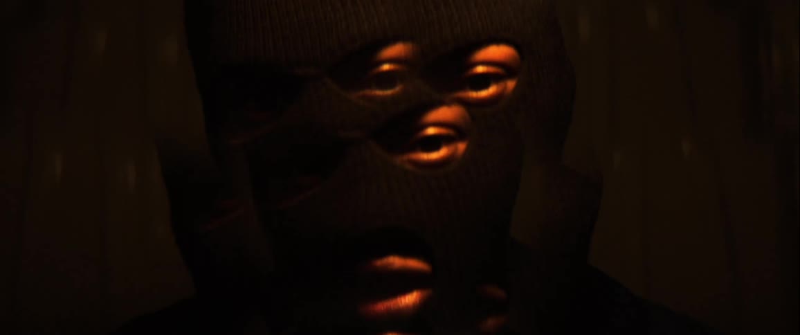 A closeup of a man wearing a black ski mask under a swinging light with his face distorted