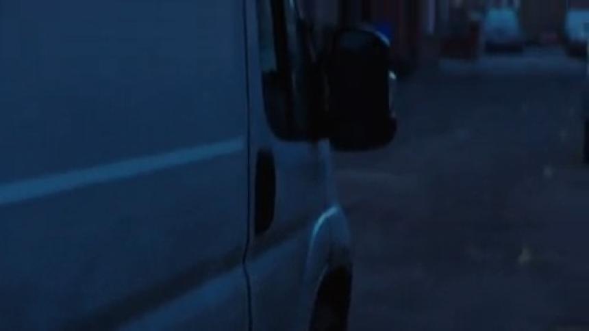 A hand holding a cigarette extends from the window of a white vehicle in a dark street scene with blurred lights and other vehicles in the background