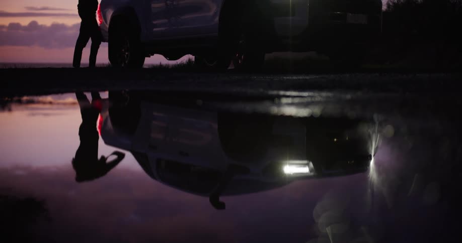 Male photographer exits a vehicle parked near a reflective body of water and gets his camera from it while red tints the sky