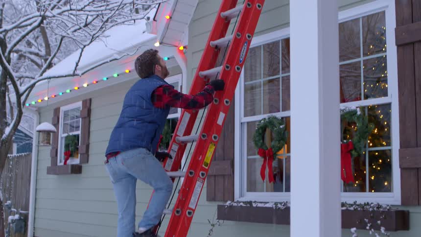 Man climbs up a ladder toward christmas lights on a snowy day
