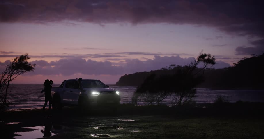 A person getting out of a pickup truck with its headlights on near the sea on a very dark morning