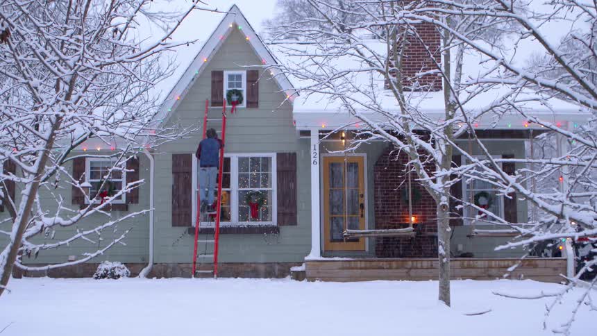 A man climbs a ladder to fix christmas lights on the facade of his house