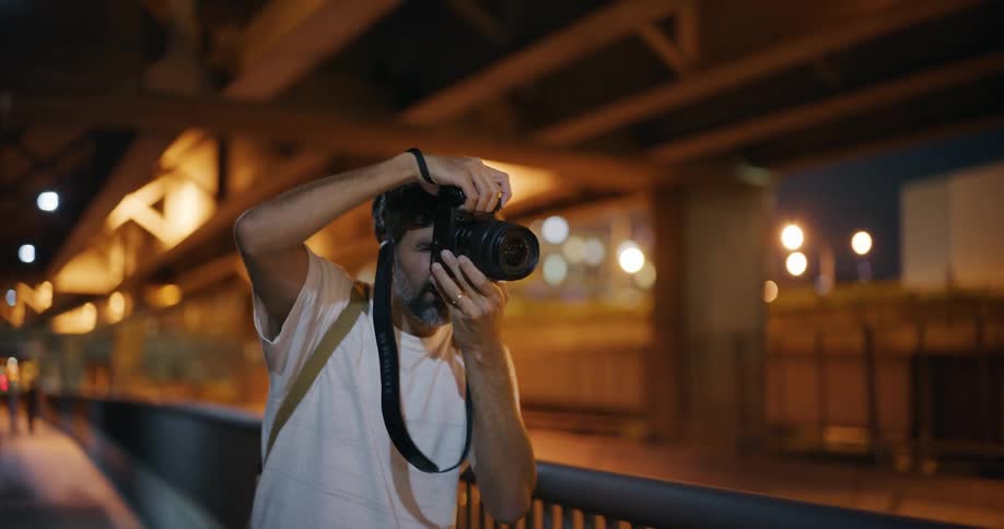 A person wearing a white shirt is taking a photograph with a camera on a bridge at night with blurred lights and structures in the background