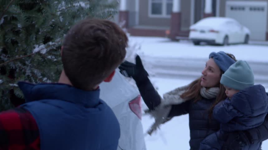 Man removing a christmas tree from the car with the help of his wife who is holding their son in her arms