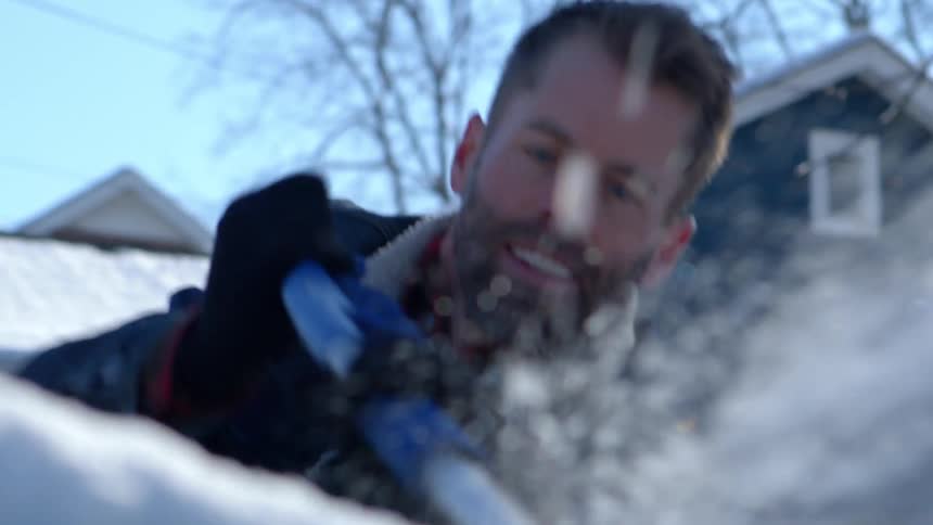 A man scrapes ice from the windshield of a car parked in a neighborhood on a sunny winter morning