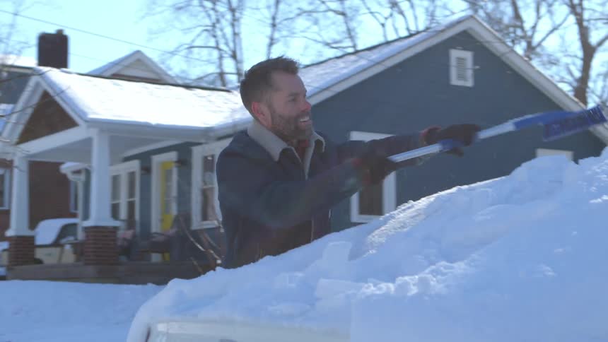 A man is clearing snow off of his car in front of a house on a sunny day