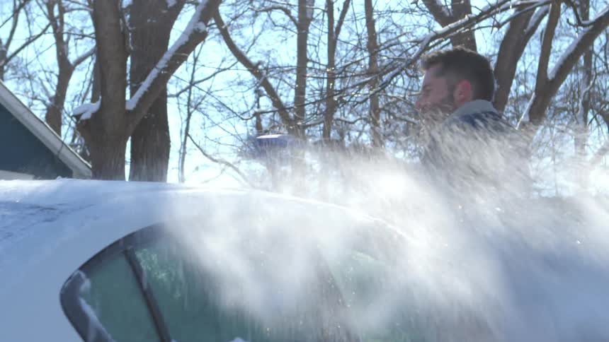 A man brushes the snow from the roof of a car parked in a neighborhood on a sunny winter morning
