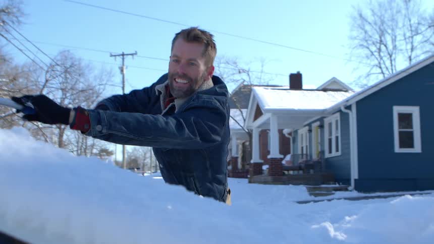 A man scrapes ice from the windshield of a car parked in a neighborhood on a sunny winter morning