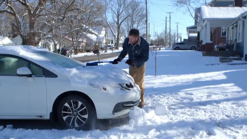 Man with a broom removing snow from the front of a white car