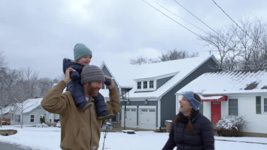 A family walks down a snowy street with one adult carrying a child on their shoulders