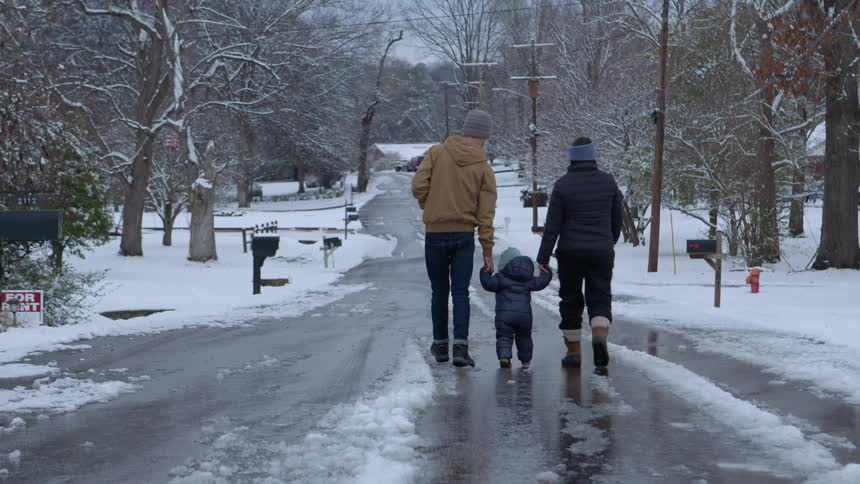 An adult man and woman are holding hands with a little baby who is walking on a street that is covered in ice and snow and they are all wearing winter clothes