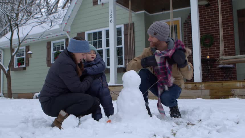 A family builds a snowman in the front yard of their snow covered house