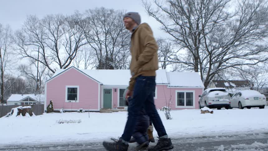 Parents walking hand in hand with their young child on a snowcovered street with a pink house in the background