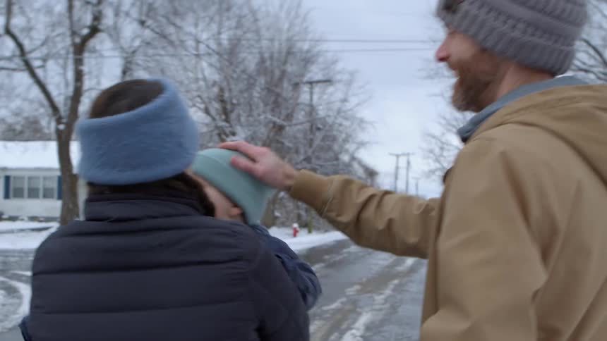 A family bundled in winter wear walks along a snow covered road with the mother holding a child in a blue hat and the father smiling beside them