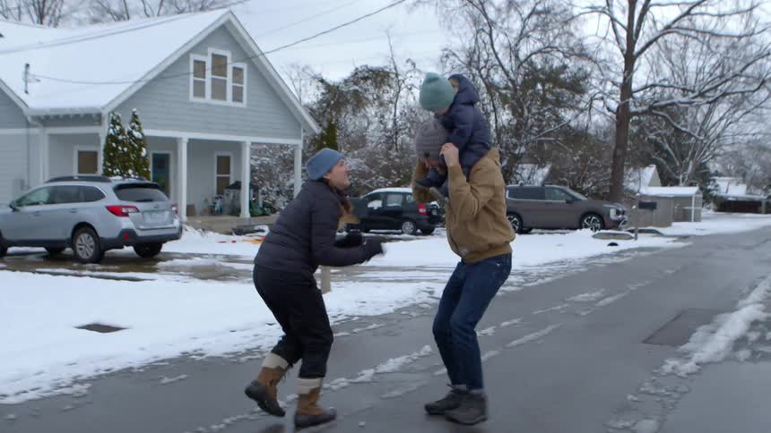 Father carries his child on his shoulders while the family walks along a residential street on a winter day