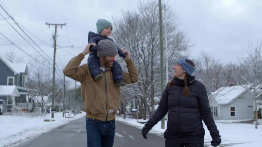 A family walks down a snowy street with one adult carrying a child on their shoulders