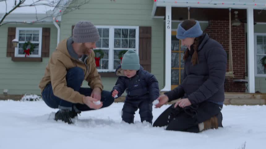 A family of a man and a woman smiling and in the middle a little boy who is playing in the snow and they are all wearing warm clothes