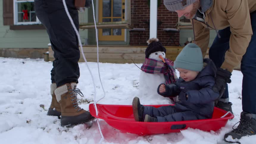 A person in a tan jacket places a child in a blue snowsuit and hat into a red sled on the snow