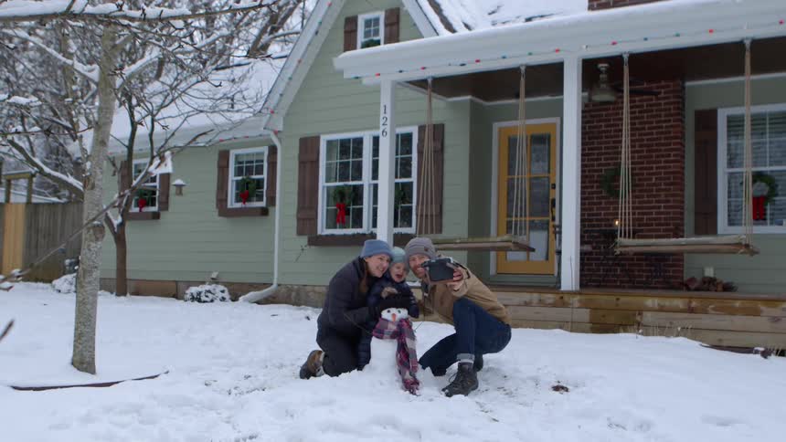 Father taking a selfie with baby and mother behind snowman built on the front lawn