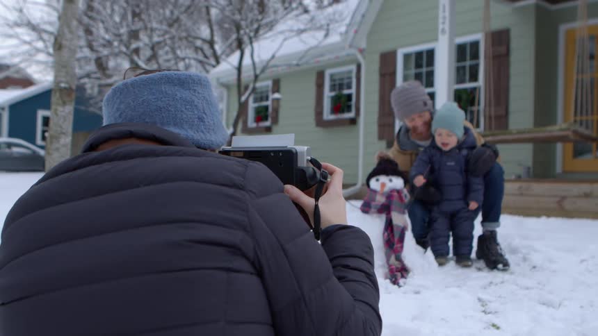 A person takes a photograph of a child and an adult standing in the snow next to a snowman in front of a house